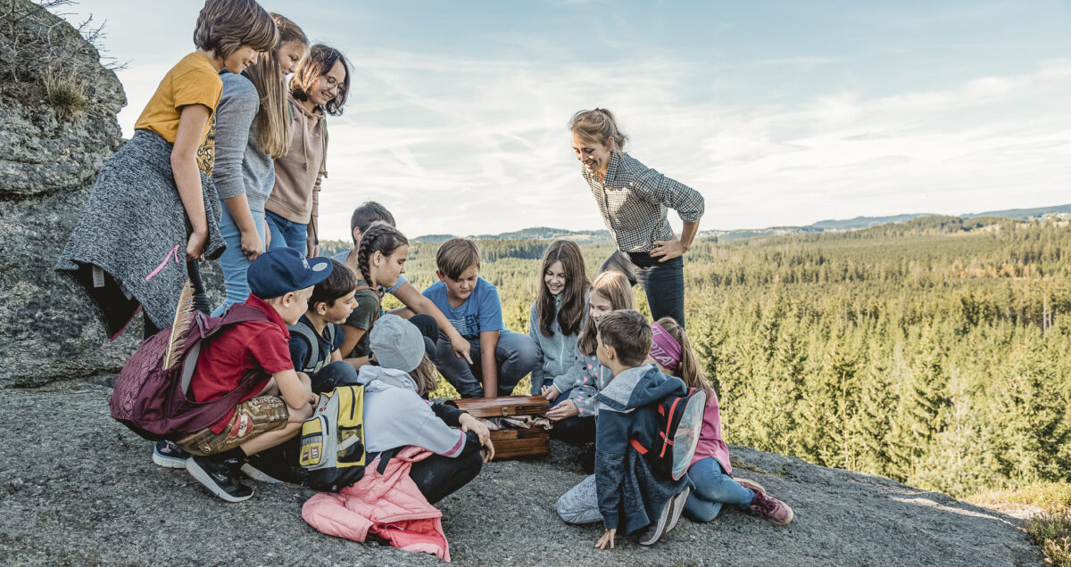 Eine Gruppe Schülerinnen und Schüler hocken und stehen im Kreis auf einer großen Steinplattform. In der Mitte der Gruppe steht ein halb geöffneter Koffer. Eine Lehrerin steht interessiert dabei.