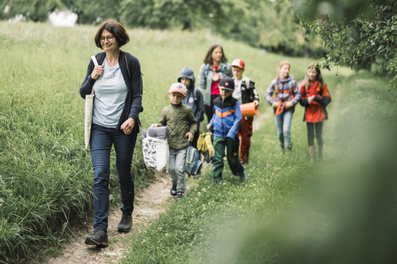 6 Volksschulkinder wandern mit 2 Betreuerinnen auf einem schmalen Weg zwischen Wiese und Bäumen