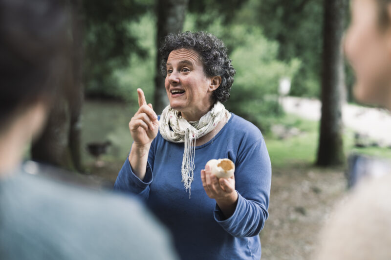 Eine Frau hat zwei aufgebrochene Eier in der Hand und spricht zu Erwachsenen, die im Vordergrund nur unscharf zu erahnen sind.