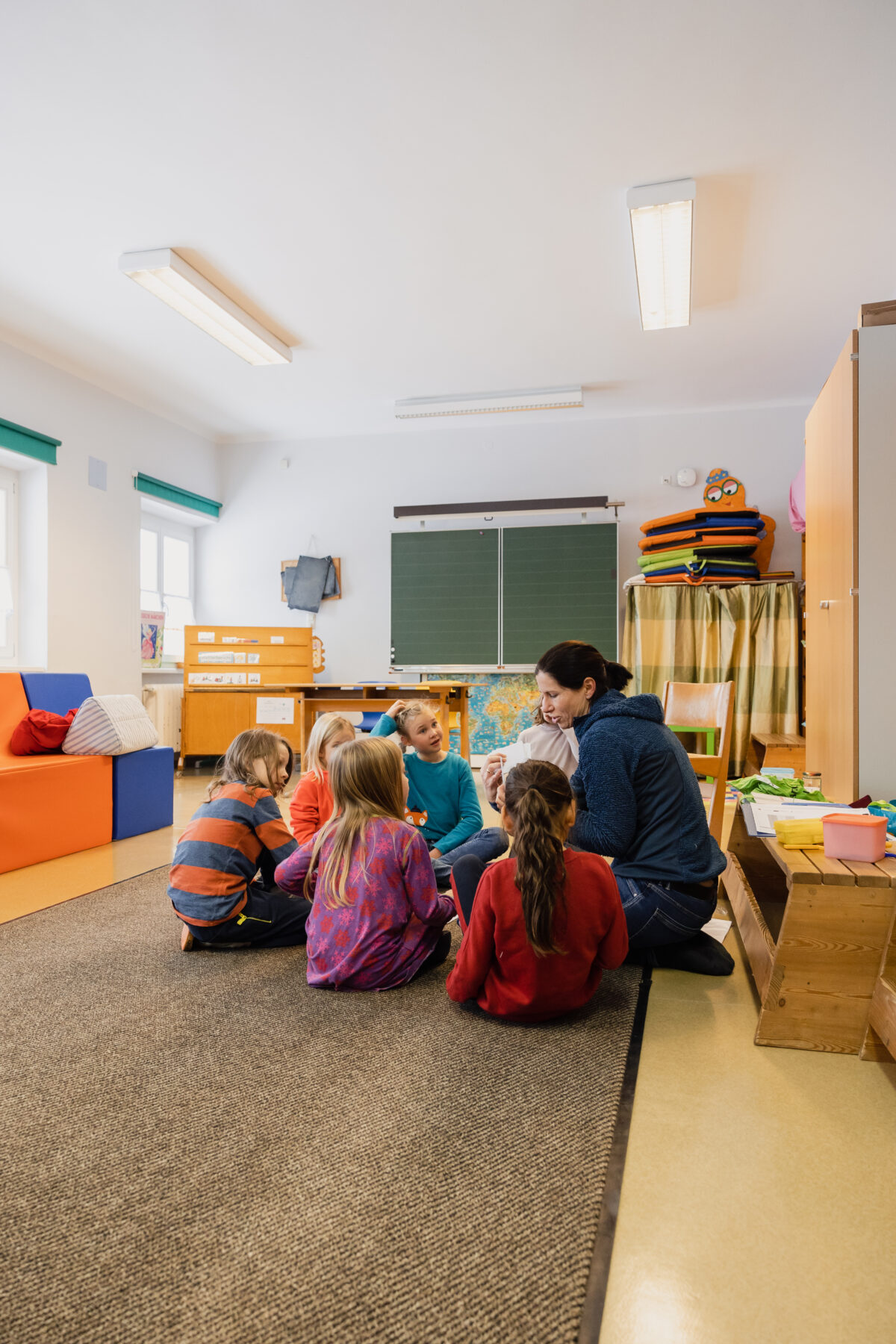 Eine erwachsene Frau spricht mit mehreren Kindern. Sie sitzen und hocken dabei am Boden einer Schulklasse.