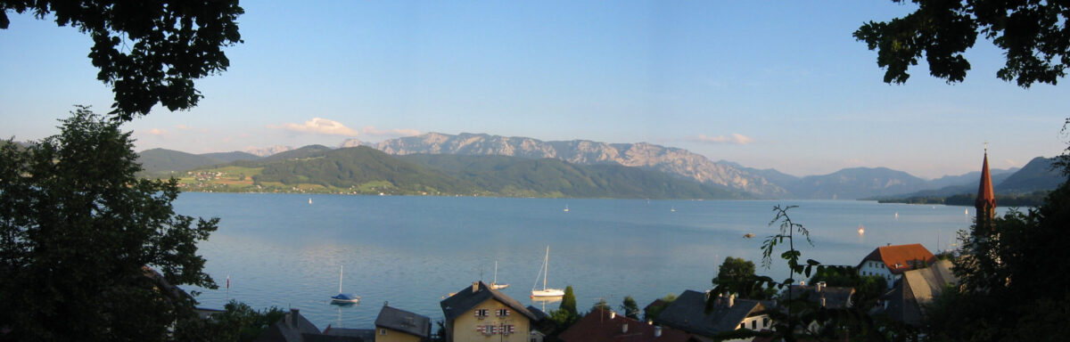 Aus höherer Lage wurde der Attersee fotografiert. Im Vordergrund die Uferstraße mit Kirchen, am anderen Ufer erhebt sich Bergmassiv
